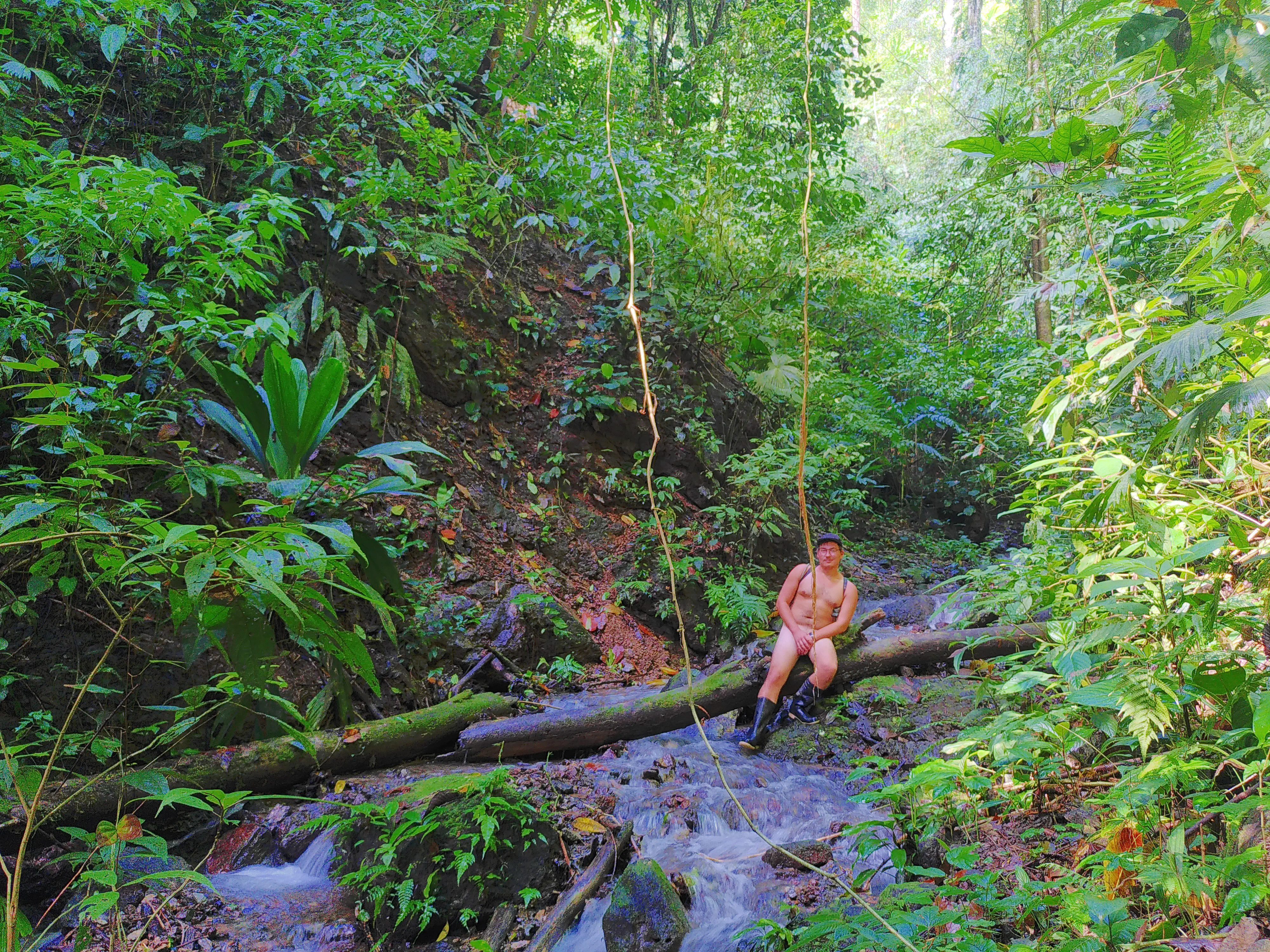 A naked man sitting on a log over a jungle river