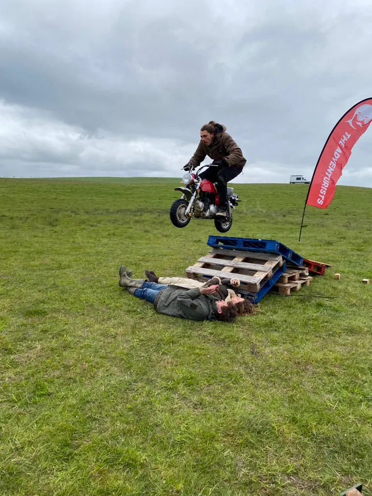 A monkey bike in mid-air, jumping over two people laying on the grass
