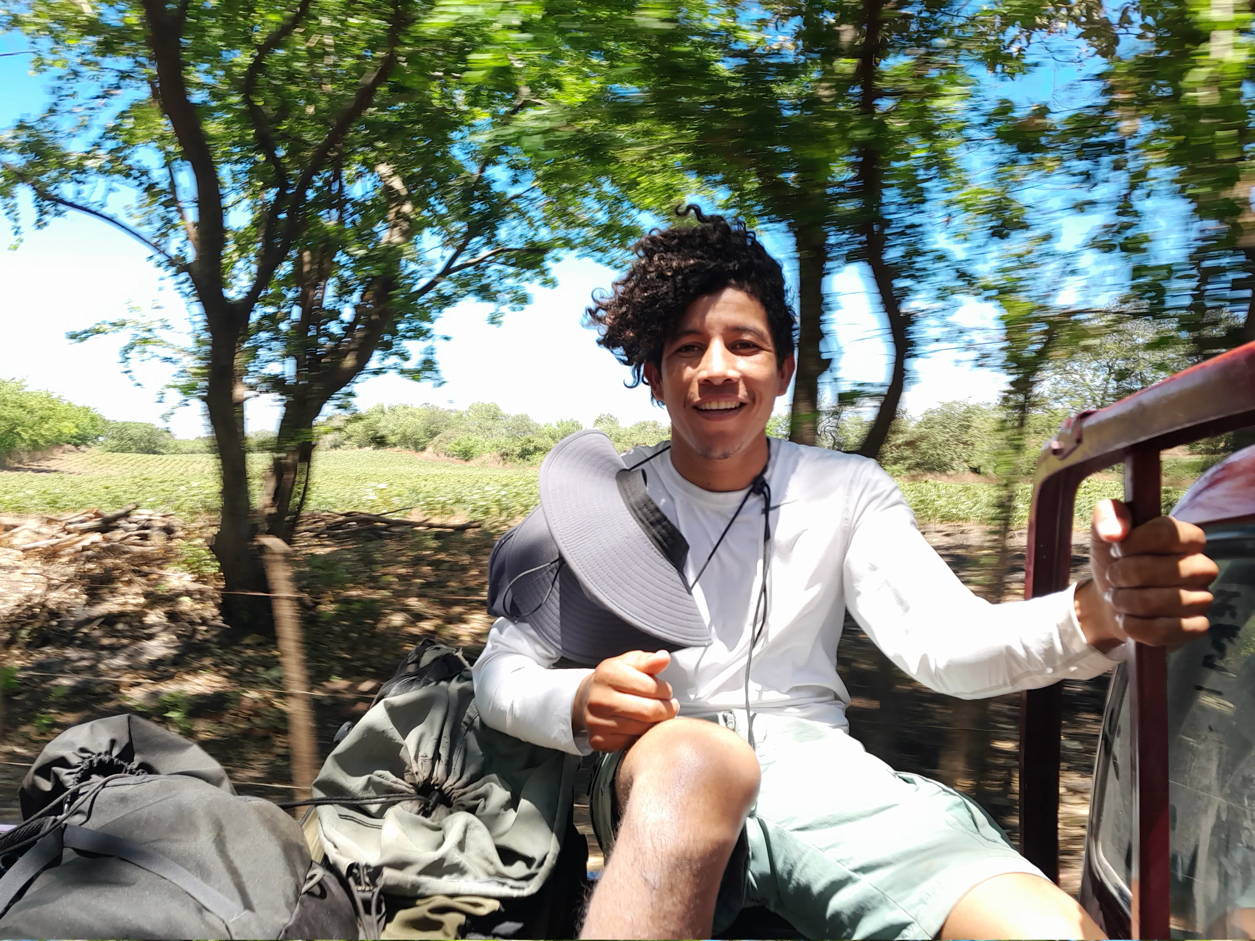 A man smiles, sat in the back of a truck as it zooms down the road
