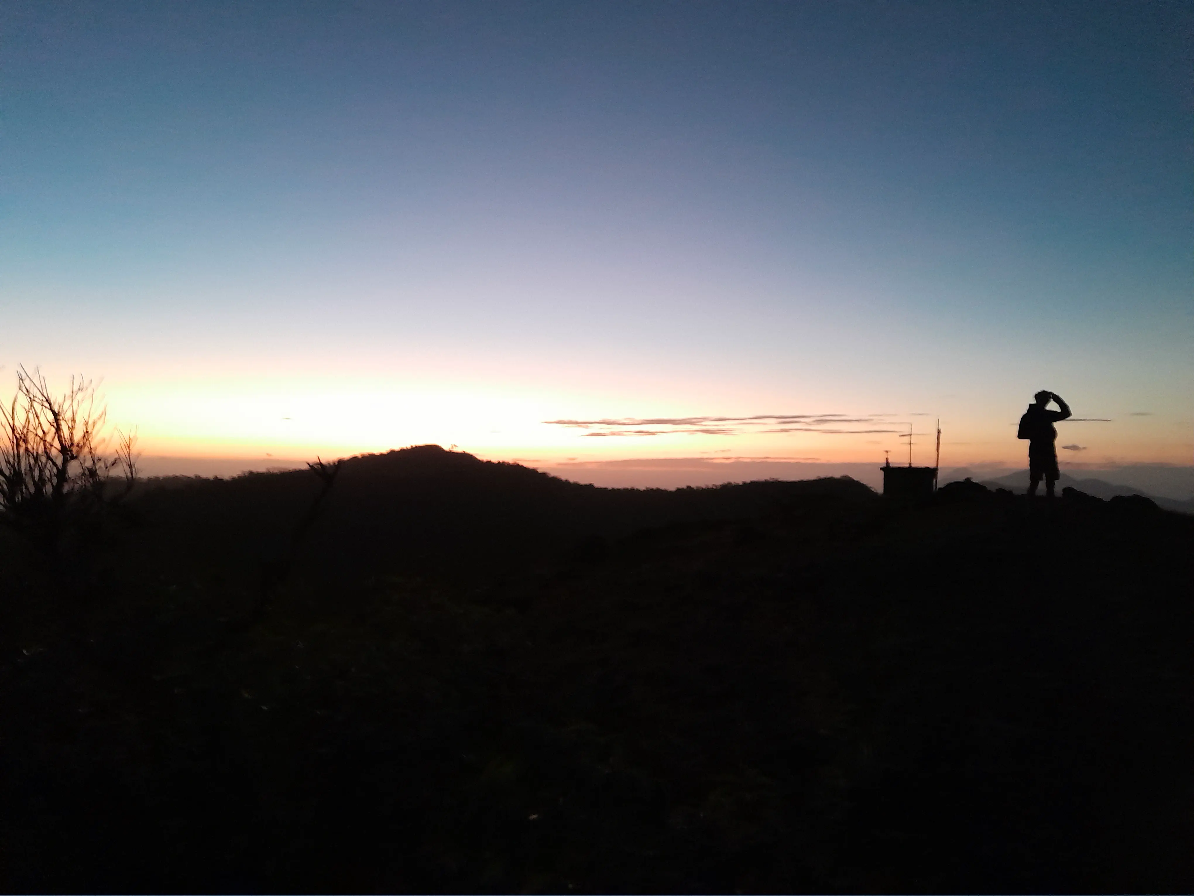 A man stands beside a small structure covered in measuring devices, both silhouetted as the sun sets