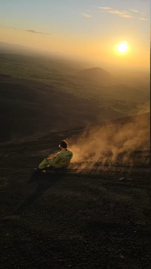 The author sliding down the side of a volcano on a wooden board as the sun sets in the background