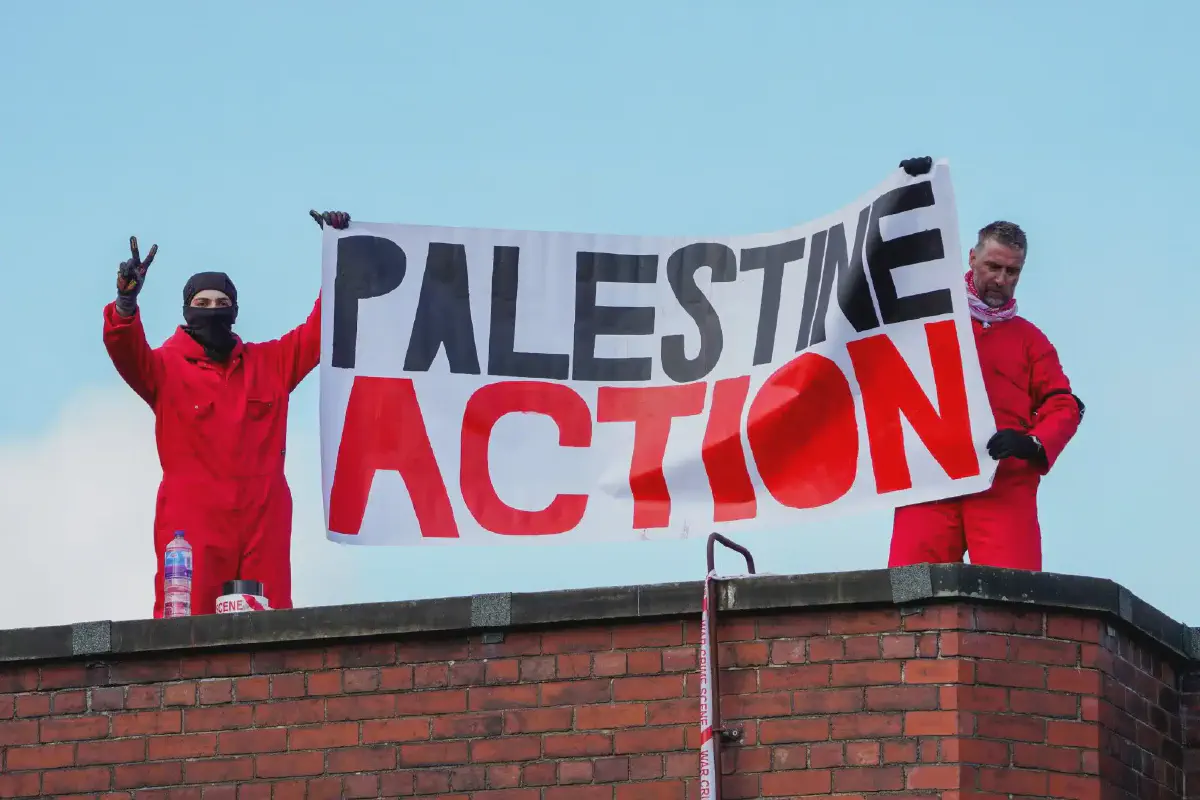 Palestine Action members in black masks and red jumpsuits protest with a banner on top of the roof of a factory