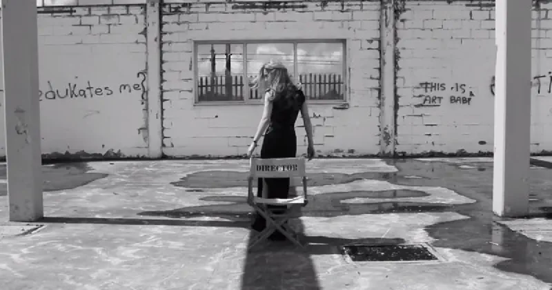 A black and white photo of a woman standing in front of a chair labelled 'Director', in a derelict building with graffiti reading 'who edukates[sic] me?' and 'this is art baby'