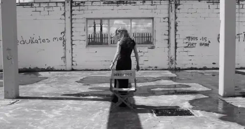 A black and white photo of a woman standing in front of a chair labelled 'Director', in a derelict building with graffiti reading 'who edukates[sic] me?' and 'this is art baby'